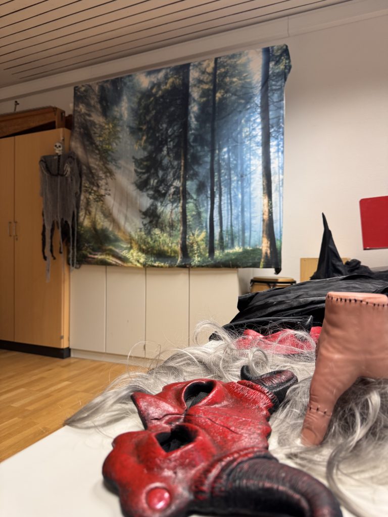 Im Hintergrund hängt eine Leinwand, die einen Wald zeigt. Im Vordergrund liegen Kostüme