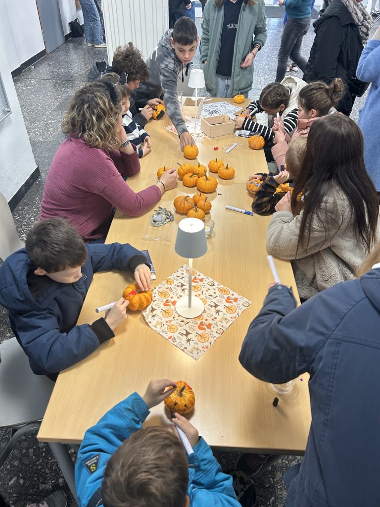 Tisch mit Minikürbissen. Um den Tisch sitzen SchülerInnen und eine Lehrkraft. Sie bemalen die Kürbisse